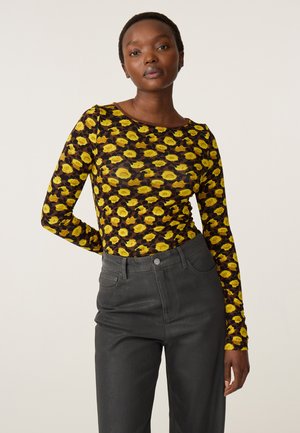 Vrouw die een geel rozenpatroon lange mouwen top en hoge zwarte broek draagt, staand met één hand achter haar rug.