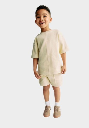 Completo a due pezzi di un beige chiaro a costine, con maniche corte e pantaloni corti. Design semplice, tessuto testurizzato, abbinato a sneakers color sabbia e calze bianche.