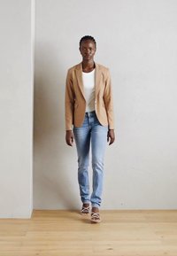 Blazer tan su una maglietta bianca, abbinato a jeans azzurri chiari. Modella indossa sandali bianchi con cinghie, in piedi su un pavimento di legno contro un muro neutro.