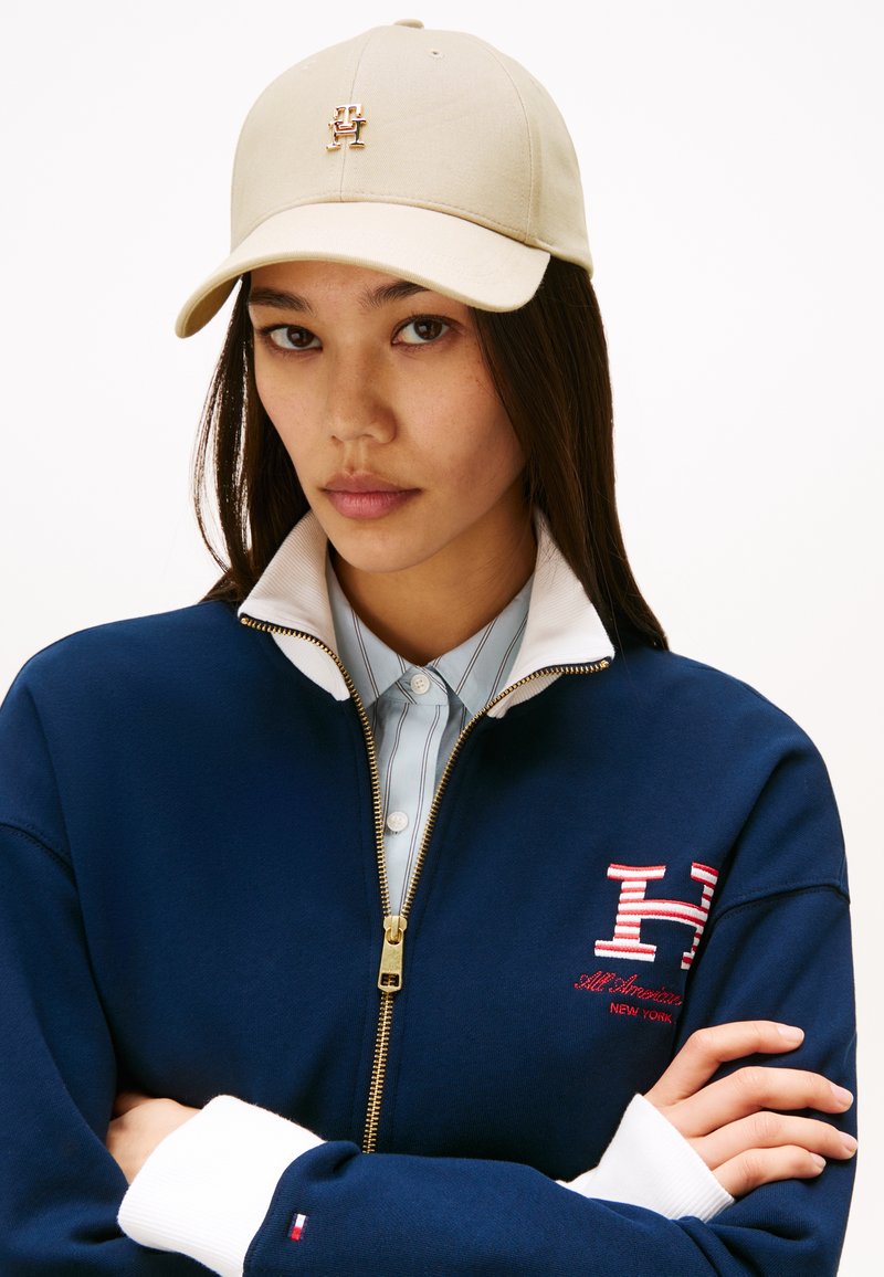 Beige baseball cap with a gold logo, worn with a navy zip-up sweater featuring a white collar and red accented details. Blue-striped shirt underneath.