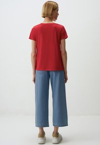 Rode top met korte mouwen en een ronde halslijn, gecombineerd met lichtblauwe wijde jeans. Lichte schoenen met een beige accent. Effen achtergrond.
