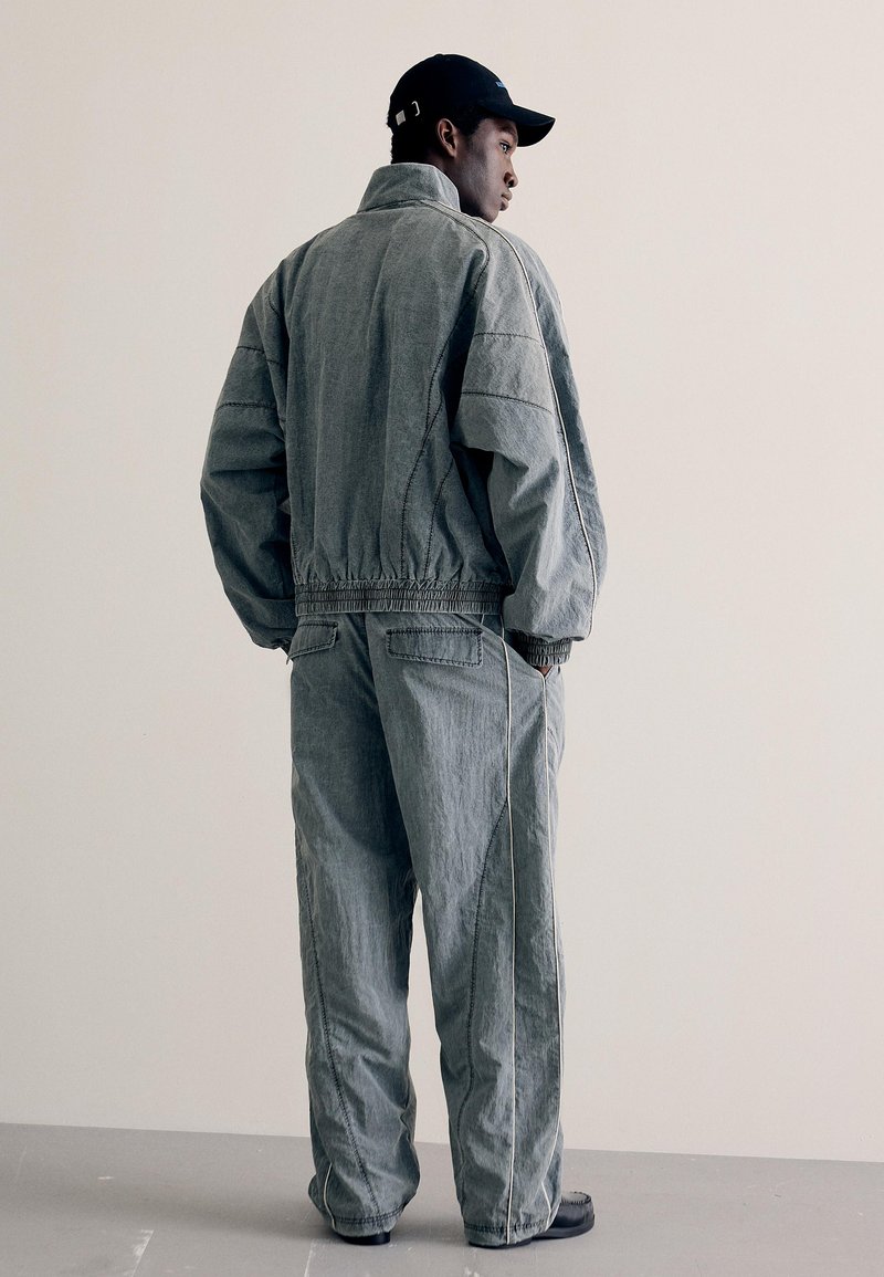Homme portant une veste en jean grise ample et un pantalon assorti, debout les mains dans les poches, de profil contre un mur uni.