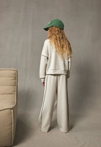 Light gray sweatshirt and wide-leg sweatpants with black side stripes, paired with a green cap, against a neutral backdrop.