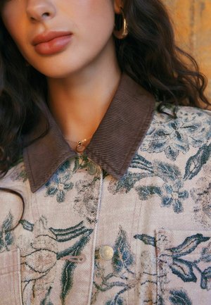 Woman with wavy dark hair wears gold hoop earring, delicate necklace, and buttoned floral jacket with dark corduroy collar.