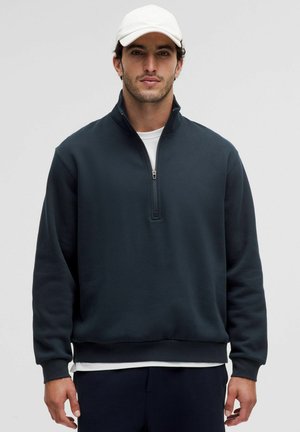 Mann trägt einen dunkelblauen Quarter-Zip-Sweatshirt, ein weißes Unterhemd, navyfarbene Hosen und eine weiße Baseballmütze vor einem schlichten Hintergrund.