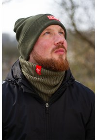 Olive green knit beanie and matching textured neck warmer with red branding, worn under a black jacket, creating a cohesive winter look.