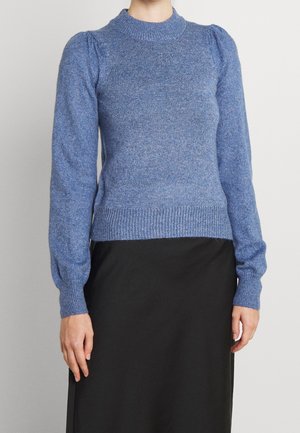 Femme portant un pull en maille bleu ajusté avec des épaules bouffantes et des manches longues, associé à une jupe noire sur un fond uni.