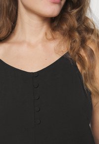 Gros plan sur le cou et la clavicule d'une femme aux cheveux bruns ondulés, portant un haut noir sans manches avec des boutons recouverts de tissu.