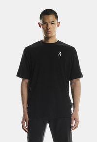 Fekete pamut póló kerek nyakkivágással és rövid ujjakkal; az elülső bal mellrészen fehér logó és az oldalon címke található.