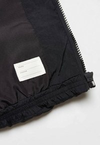 Intérieur d'une veste noire montrant une étiquette avec un nom et un numéro de téléphone cousue à l'intérieur, près d'une fermeture éclair noire partiellement zippée avec le texte "HEAT" sur le tirette.