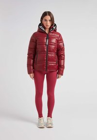 Vadderad röd jacka med en svart huva, med dragkedja fram och två sidofickor. Materialet har en glänsande yta och är kombinerat med röda leggings och sneakers.