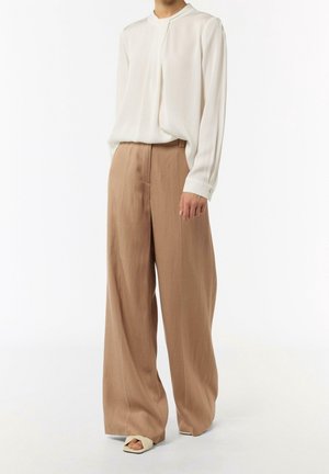 Modèle portant un pantalon beige taille haute à jambes larges, une blouse blanche à manches longues avec un détail de plis, et des sandales crème à bout ouvert sur fond blanc.