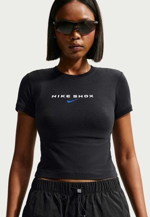 Kobieta ubrana w czarny krótki t-shirt Nike Shox, czarne sportowe spodenki i ciemne okulary przeciwsłoneczne typu wraparound, zwrócona przodem na jasnym tle.