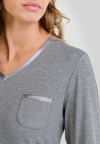 Mujer con una camiseta de manga larga gris, con escote en V y un pequeño bolsillo decorado con satén en el pecho.