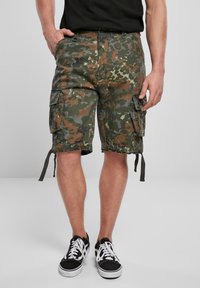 Bermudy w kamuflażowym wzorze, z wieloma kieszeniami, luźnym krojem, ściągaczami na dole i wykonane z wytrzymałego materiału. Nosi się je z czarnymi sneakerami.