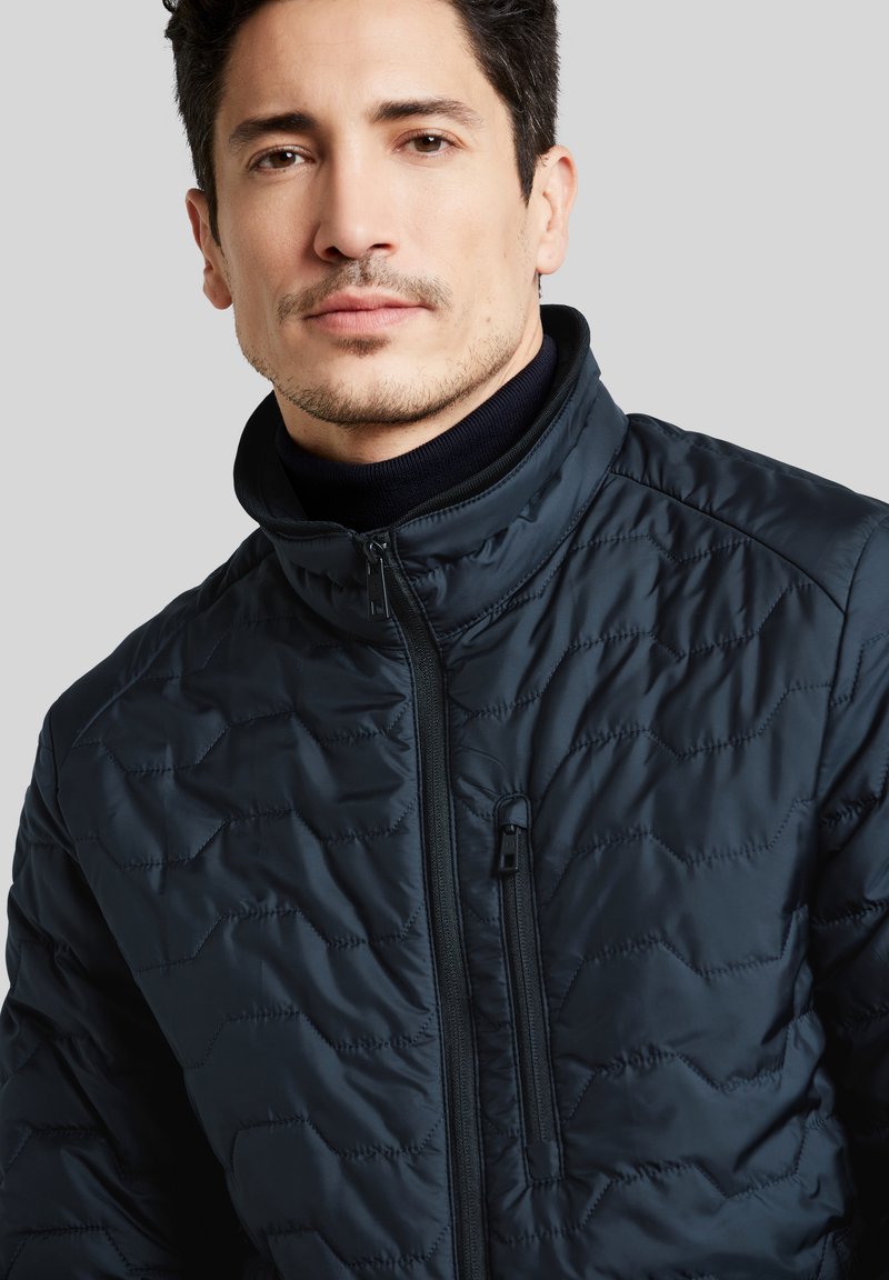 Veste rembourrée marine avec un col haut, présentant un motif matelassé, fermeture zippée à l'avant et poche zippée sur le côté pour le rangement. Fini lisse et brillant.