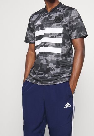 Man wearing black and gray camo-patterned Adidas shirt with white stripes, paired with blue Adidas pants with white side stripes.
