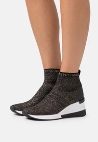 Bottines noires en tricot avec des fibres métalliques, semelle blanche et talon structuré. Présente une bande logo en haut pour un détail.