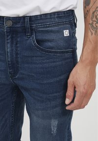 Jeans in denim blu scuro con una vestibilità slim, caratterizzati da una tasca frontale per le monete, leggere sfumature e un piccolo etichetta con il logo sulla vita.