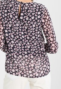 Blouse à fleurs avec un fond bleu marine orné de petites fleurs roses et blanches, manches longues en tissu transparent et détail fente au dos.