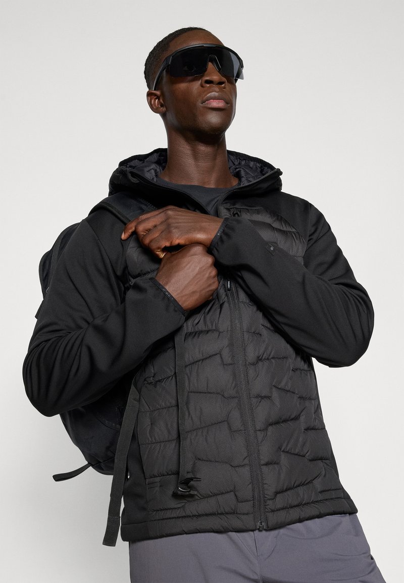 Czarna kurtka puchowa o teksturowanym wykończeniu, z kapturem i zamkiem, zestawiona z czarną koszulką z długim rękawem oraz plecakiem. Ciemne okulary przeciwsłoneczne.