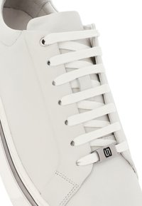 Sneaker bianco con lacci bianchi passati attraverso occhielli metallici, dotato di un piccolo disco d'argento con un logo nero vicino ai lacci inferiori.