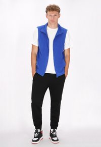 Blaues Fleece-Weste mit Reißverschluss, getragen über einem weißen T-Shirt. Kombiniert mit schwarzen Hosen und schwarz-roten High-Top-Sneakern.
