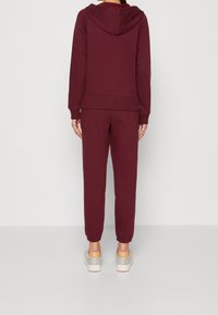 Set di felpa e pantaloni jogger bordeaux, realizzato in tessuto morbido. La felpa presenta un cappuccio con cordino e una tasca a marsupio; i pantaloni jogger hanno polsini elasticizzati.