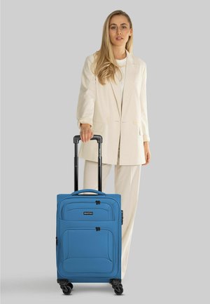 Donna in completo beige in piedi che tiene la maniglia di una valigia blu con ruote e maniglia telescopica su sfondo grigio.