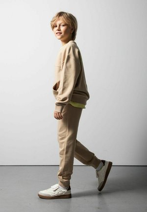 Blond dziecko w beżowej bluzie i spodniach, białe trampki, spacerujące wewnątrz przed jednolitą jasnoszarą ścianą.