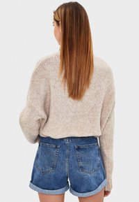 Mujer con cabello largo y liso lleva un suéter de punto beige y pantalones cortos de mezclilla azul arremangados, vista desde atrás contra un fondo simple.