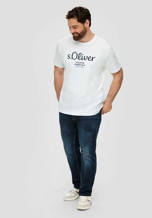 Mann mit Bart, der ein weißes "s.Oliver"-T-Shirt, dunkelblaue Jeans und weiße Turnschuhe trägt, steht mit den Händen in den Taschen, schaut nach unten und lächelt.