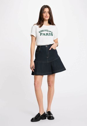Giovane donna che indossa una t-shirt bianca con scritta "Couture Paris", gonna in denim scuro e mocassini neri, in piedi con una mano in tasca.