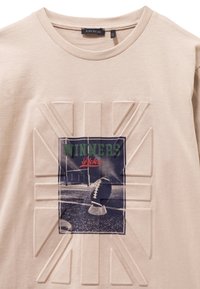 Beige t-shirt van katoen, met een verhoogd ontwerp en een voetbalafbeelding met de tekst "Winners" in groen en rood, gecentreerd op de voorkant.