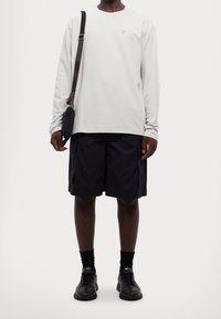 Helles graues Langarmhemd mit lila Logo, schwarze Cargo-Shorts mit Seitentaschen, schwarze Sneakers und eine kleine schwarze Umhängetasche.