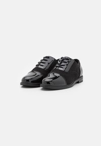 Chaussures en cuir noir avec un embout en vernis brillant et une tige en daim, comportant six œillets et une semelle plate. Texture lisse avec un design classique.