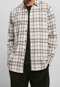 Man in een wit, rood en zwart geruit overhemd over een zwart T-shirt met zwarte broek, handen in de zakken.