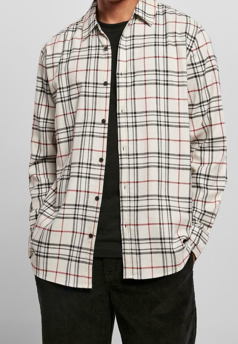 Man in een wit, rood en zwart geruit overhemd over een zwart T-shirt met zwarte broek, handen in de zakken.