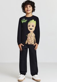 Maglietta nera a maniche lunghe con una grande grafica di Groot e accenti verdi, abbinata a pantaloni neri e sneakers bianche.