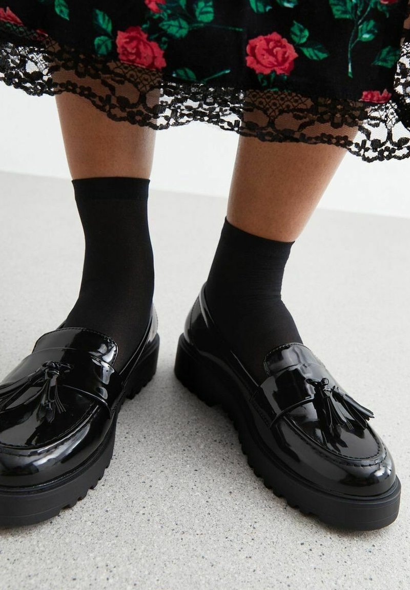 Mocassins à glands noirs brillants portés avec des chaussettes noires et une jupe noire à motifs floraux avec des roses rouges et une bordure en dentelle au-dessus des chevilles.