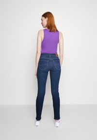 Top sin mangas morado combinado con jeans de mezclilla azul oscuro; los jeans tienen bolsillos traseros y un parche de cuero en la cintura. Textura suave.