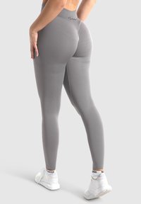Szare legginsy z wysokim stanem, bezszwowym wzornictwem i gładką fakturą. Noszone z białymi butami sportowymi, z logo na pasie.