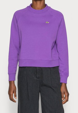 Persoon draagt een paarse trui met lange mouwen en een ronde hals, met een klein groen krokodillenlogo op de linkerborst en donkere broek.