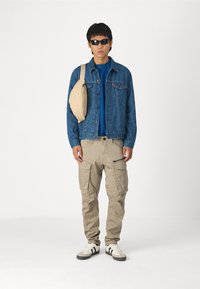 Modelo vistiendo una chaqueta de mezclilla azul, camisa azul marino, pantalones cargo beig y gafas de sol, con un bolso cruzado color beige y zapatillas blancas con rayas negras.