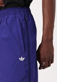 Zbliżenie osoby noszącej fioletowe spodnie dresowe Adidas z elastycznym pasem, bocznymi kieszeniami i białym logo Adidas na udzie.