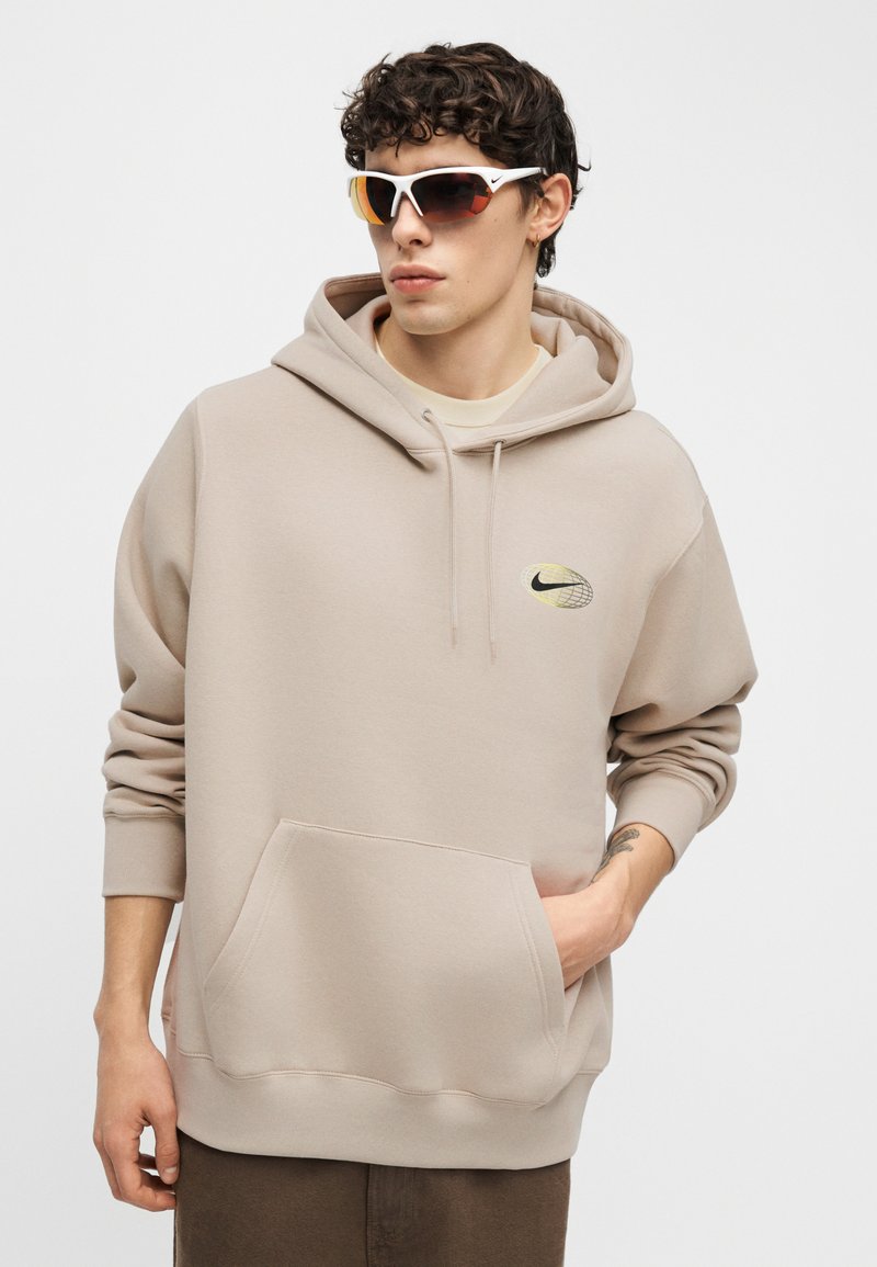 Giovane uomo con felpa beige Nike e occhiali da sole sportivi bianchi, con la mano destra nella tasca frontale, in piedi davanti a uno sfondo semplice.