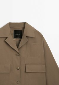 Veste marron clair à boutons avec un col, trois boutons, et trois poches avant, dont une poche poitrine et deux poches à rabat.