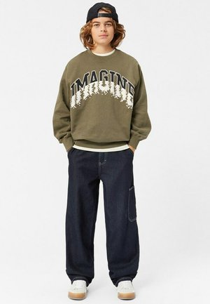 Olivgrön sweatshirt med "IMAGINE" grafik, kombinerad med mörkblå vida jeans med sidofickor. Vita sneakers fulländar looken.