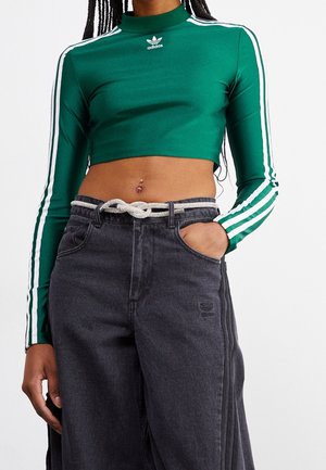 Person trägt grünes, langärmeliges Adidas-Cropped-Shirt mit weißen Streifen und schwarze High-Waist-Jeans mit einem geknoteten Seilgürtel, Hand in der Tasche.