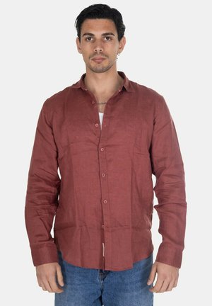 Uomo con capelli corti e scuri che indossa una camicia color ruggine abbottonata sopra una maglietta bianca e jeans blu, in piedi davanti a uno sfondo semplice.